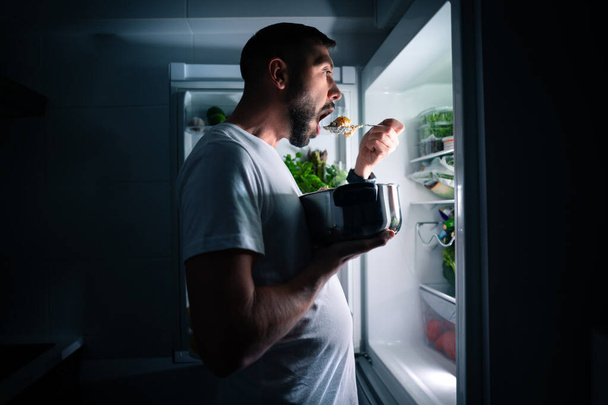 Uomo affamato che mangia cibo di notte dal frigo aperto. Uomo che prende spuntino di mezzanotte dal frigorifero - Foto, immagini