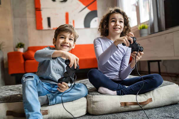 Zwei Kinder kleine kaukasische Bruder und Schwester glückliche Kinder Geschwister Jungen und Mädchen spielen Videospielkonsole mit Steuerknüppel oder Controller, während sie zu Hause sitzen echte Menschen Familienfreizeitkonzept - Foto, Bild