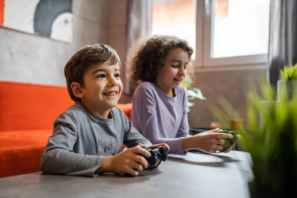 Two children small caucasian brother and sister happy children siblings boy and girl playing video game console using joystick or controller while sitting at home real people family leisure concept - Photo, Image