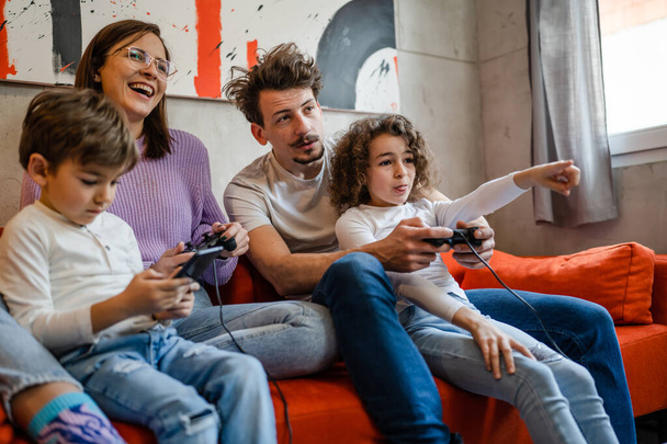 csoport emberek kaukázusi család férfi és nő anya és apa gyerekekkel fiú és lány ül a kanapén otthon videojáték konzol segítségével joystick vagy vezérlő valós emberek koncepció - Fotó, kép
