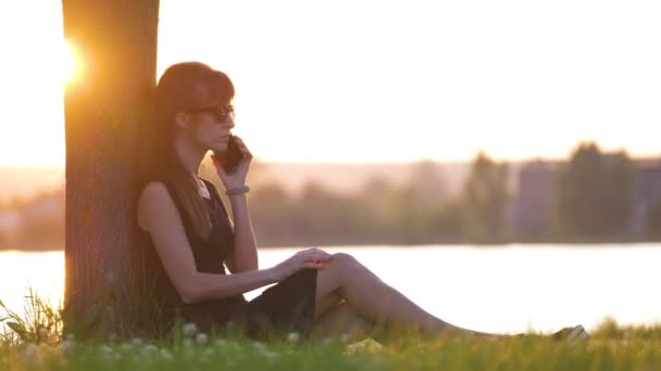 Mujer joven y feliz relajarse en el parque de verano césped junto al lago hablando en su teléfono móvil al aire libre en la noche cálida. Concepto de comunicación y conexión móvil - Metraje, vídeo
