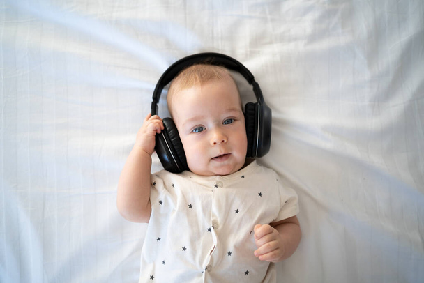 vista superior, niño escuchando música en auriculares inalámbricos, oídos de niños con auriculares, niño pequeño en la cama, fondo blanco - Foto, imagen