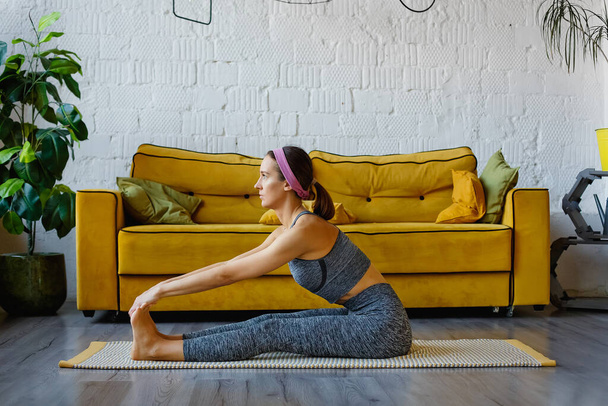 Eine sportliche junge Frau, die morgens allein im Wohnzimmer neben dem gelben Sofa Übungen macht, ein ernstzunehmendes Mädchen in Sportkleidung, Yoga-Training zu Hause für das Konzept eines gesunden Lebensstils, Seitenansicht. - Foto, Bild