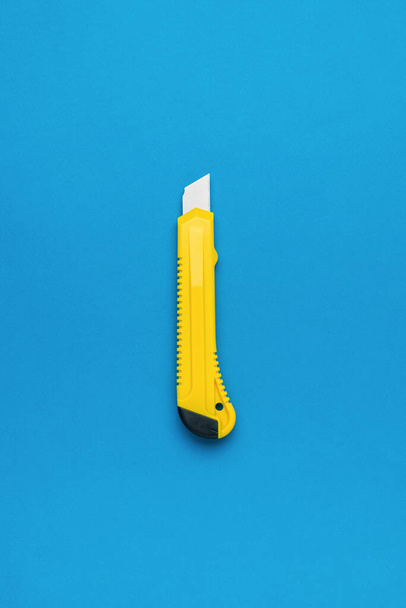 Foto de um design minimalista de uma faca de papelaria amarela sobre um fundo azul. A composição central da faca de papel. Depósito plano. - Foto, Imagem