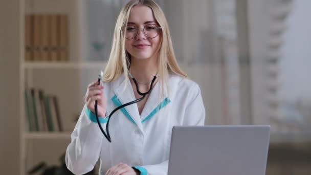 Médica branca pediatra cardiologista mulher enfermeira médica especialista em óculos e vestido olhando para a câmera sorrindo sentado à mesa com laptop na clínica do hospital com estetoscópio - Filmagem, Vídeo