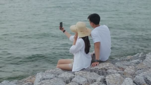 Las parejas felices usando un teléfono inteligente toman una foto y selfie mientras se sientan en la piedra en la playa durante el hermoso atardecer. Pareja romántica viajan en fin de semana de vacaciones. - Imágenes, Vídeo