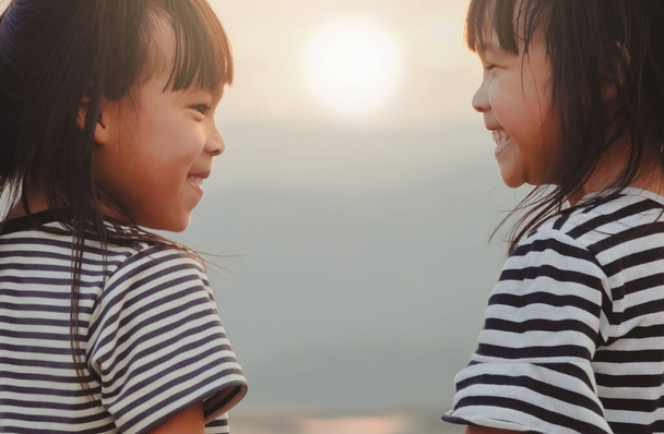Nette kleine Schwestern, die einander anschauen und vor dem Hintergrund des Sonnenuntergangs lächeln. Das Konzept der Familie und Geschwisterliebe - Foto, Bild