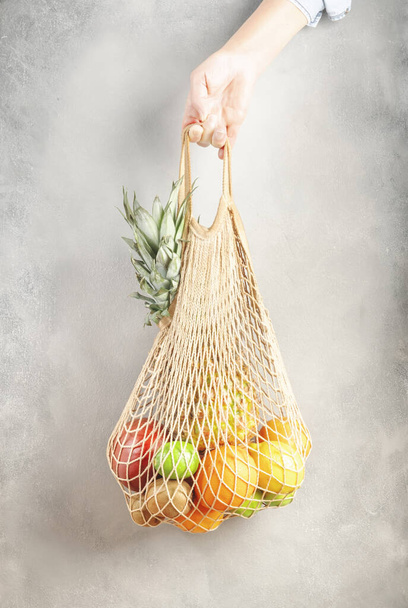 Woman hand holding eco friendly shopping bag with various tropical fruits on gray background. Sustainable lifestyle. Plastic free. Zero waste concept - Photo, Image