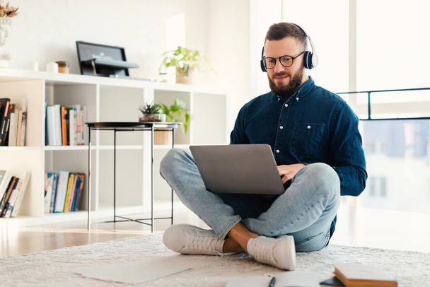 Мужчина фрилансер просматривает Интернет с помощью ноутбука носить наушники, сидя в помещении - Фото, изображение