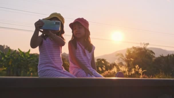 Zwei Schwestern sitzen auf den Gleisen und machen bei Sonnenuntergang Selfies mit ihren Smartphones. Asiatische Schwestern genießen die gemeinsame Zeit im Urlaub. - Filmmaterial, Video