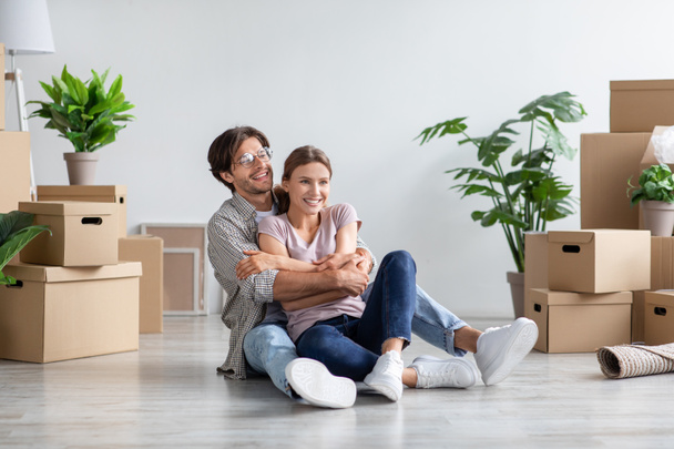 Glad millennial caucasian male in glasses and female in casual hugs, sits on floor among cardboard boxes - Foto, imagen