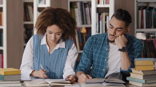 Két diák srác és lány ül az íróasztal osztályban jegyzeteket ír jegyzetfüzetbe információt a tankönyvből olvasás könyv fiatal férfi diák elalszik a leckében hallgatás tanár unalmas előadás - Felvétel, videó