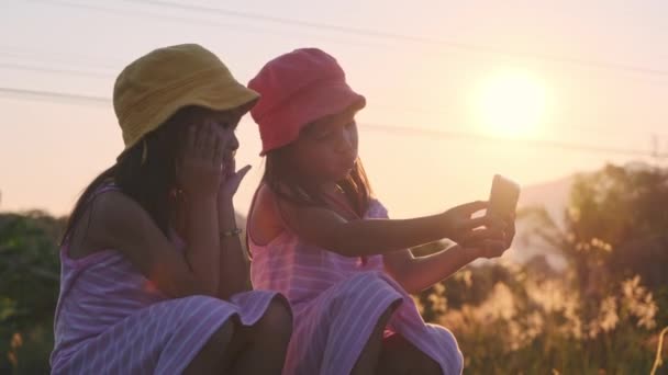 Due sorelle si siedono sui binari e scattano selfie insieme ai loro smartphone al tramonto. Asiatico sorelle godere di spendere tempo insieme in vacanza. - Filmati, video