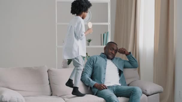 Mature african american exhausted tired after work father sitting on sofa little cheerful noisy daughter jumping on couch interferes disturbs dad from resting man upset by behavior child kid girl play - Πλάνα, βίντεο