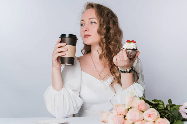 Μια όμορφη νεαρή γυναίκα με μπούκλες σε λευκό φόντο κρατά ένα κεκάκι με κρέμα και μούρα και ένα πλαστικό φλιτζάνι καφέ ή τσάι στο χέρι της. Πίνεις ένα ποτό με κέικ. - Φωτογραφία, εικόνα