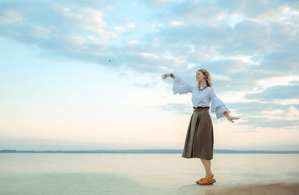 Portrait of young european woman standing, smiling and dancing on sea landscape background. Good morning mood. Nature inspiration. Beautiful girl in white blouse and khaki skirt. Travel and vacation. - Photo, image