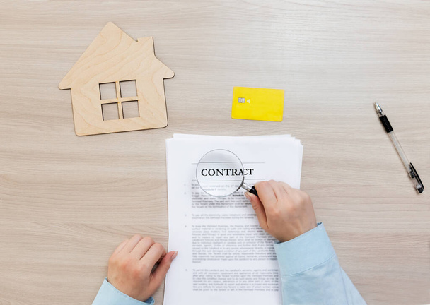 Studying a contract for the purchase or lease of real estate before signing. Women's hands hold a magnifying glass and study the contract. A document, a wooden house symbol, a bank card and a pen lie next to a contract on a wooden table. - 写真・画像