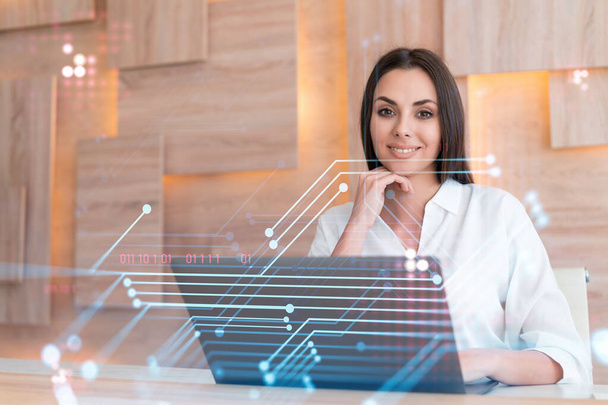 Attractive businesswoman in white shirt at workplace working with laptop to optimize development by implying new technologies in business process. Hi tech hologram over office background - Foto, afbeelding