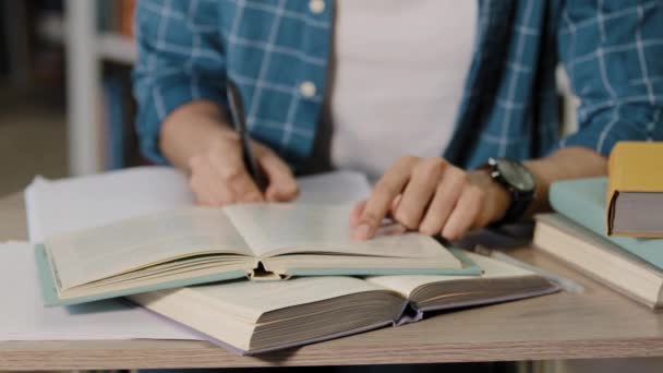 Κοντινό πλάνο αρσενικά χέρια αγνώριστη νεαρός φοιτητής κάθεται στη βιβλιοθήκη κάνει κατ 'οίκον προετοιμασία τεστ σημειώσεις γραφής στο σημειωματάριο από το βιβλίο διαβάζει προσεκτικά τις πληροφορίες στο βιβλίο γράφει σε χαρτί - Πλάνα, βίντεο