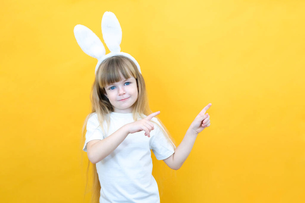 cheerful girl with rabbit ears on her head on a yellow background. Funny happy child points fingers at an empty space. copy space for text, mockup - Photo, image