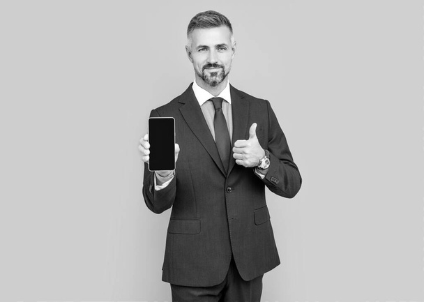 Reifer Mann Influencer im Anzug präsentiert Handy-Gerät und zeigt Daumen hoch, bestes Angebot. - Foto, Bild
