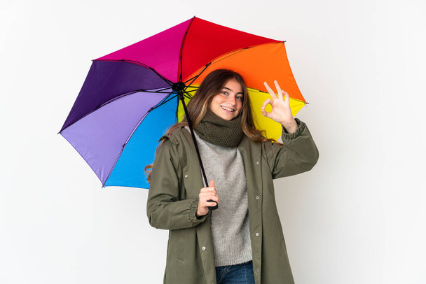 Mujer caucásica joven sosteniendo un paraguas aislado sobre fondo blanco mostrando signo de ok con los dedos - Foto, Imagen