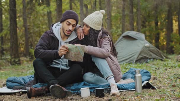 幸せな若いインドのカップルのハイカーは、テントの近くに自然の中で紙の地図を見て座ってルートの方向をチェックするガイドブックを使用して2人の旅行者を愛するロマンチックな休暇旅行の観光コンセプトとバックパック - 映像、動画