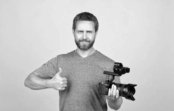Neşeli sakallı adam kameramanla video bloğu için film çekiyor, başparmağını gösteriyor, filme çekiyor. - Fotoğraf, Görsel