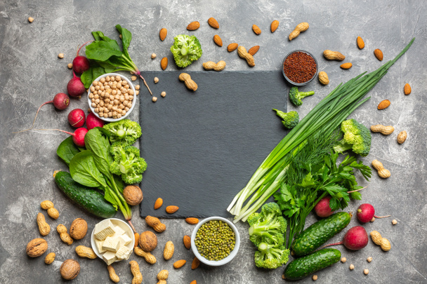 Eine Proteinquelle für Vegetarier. Gesundes, sauberes Essen: Grünzeug, Gemüse, Nüsse und Hülsenfrüchte von oben auf einem Betongrund mit einem schwarzen Schneidestein in der Mitte. - Foto, Bild