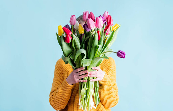 International Women's Day. pretty charming brunette young woman dressed yellow clothes holding tulip bunch isolated blue color background - Фото, изображение