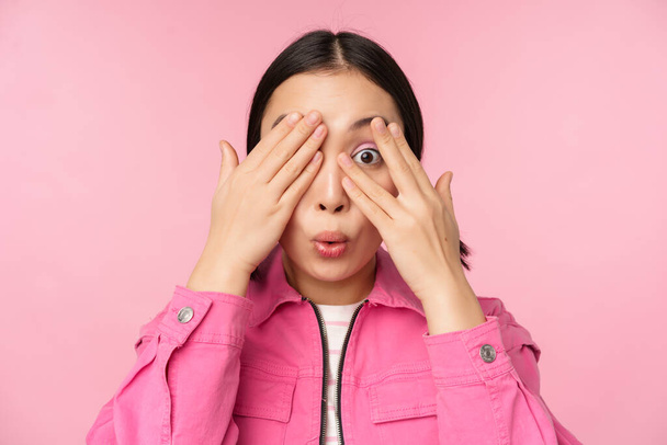 Крупним планом портрет молодої азіатки виглядає здивованою, вираженою здивуванням і дивом, дивлячись крізь пальці, стоїть над рожевим тлом
 - Фото, зображення