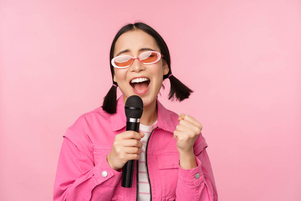Счастливая красивая девушка, поющая с микрофоном, наслаждающаяся караоке, позирующая на розовом студийном фоне - Фото, изображение