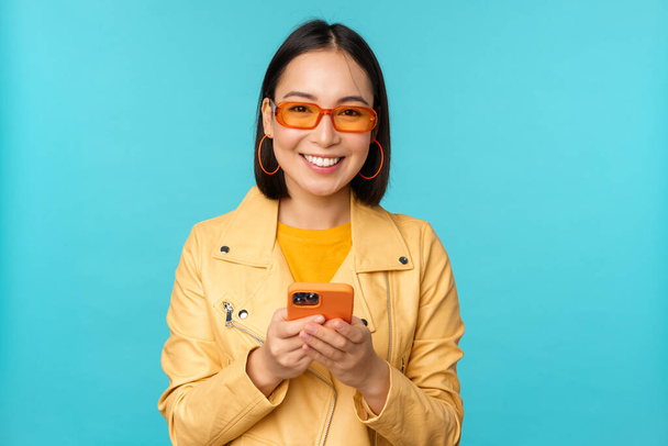Lächelndes asiatisches Mädchen mit Sonnenbrille, Smartphone-App, Handy in der Hand, vor blauem Hintergrund stehend - Foto, Bild