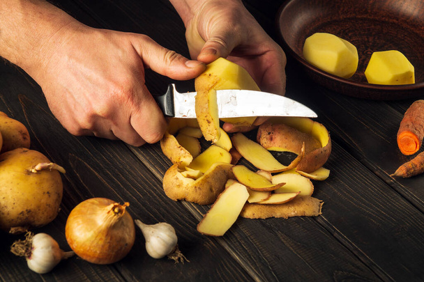 Mãos de cozinheiro close-up estão descascando batatas cruas com uma faca para preparar um prato nacional ou batatas fritas. Delicioso café da manhã ou almoço vegetal. - Foto, Imagem