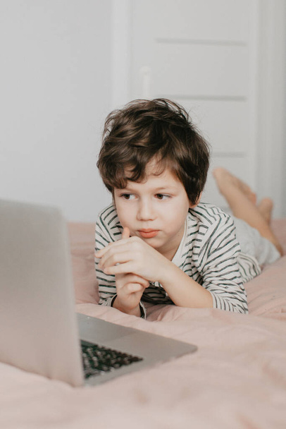 Mały chłopiec leży w sypialni na łóżku i patrzy na laptopa. - Zdjęcie, obraz