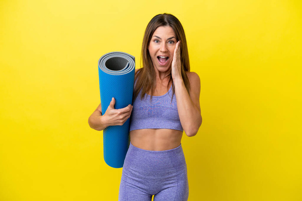 Sport woman going to yoga classes while holding a mat isolated non yellow background with surprise and shocked facial expression - Photo, image