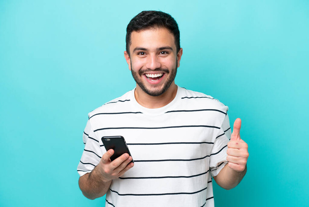 Young Brazilian man isolated on blue background using mobile phone while doing thumbs up - Photo, Image