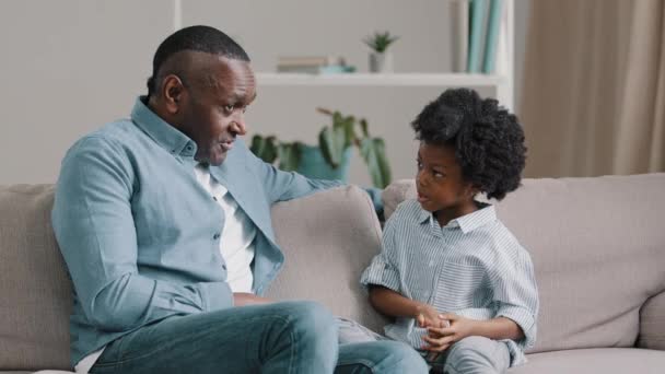 Зрілий афро-американський чоловік, який розмовляє з маленькою дівчинкою, яка сидить на дивані в кімнаті з люблячим турботливим батьком, запитує маленьку дочку, як вона провела день у школі, відпочиваючи разом, приємну родинну розмову. - Кадри, відео