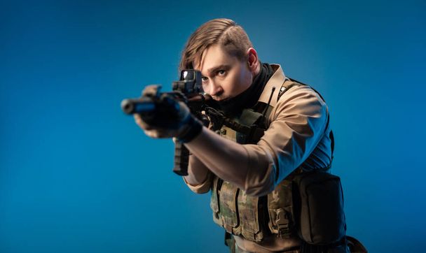 un soldado agente especial masculino en un chaleco antibalas con un rifle automático sobre un fondo azul - Foto, Imagen