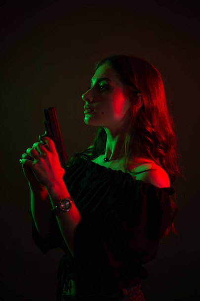 A beautiful young woman in the dark in a club holds a gun in her hands. silhouette of a girl in red-green colors. Lantern light - Foto, immagini
