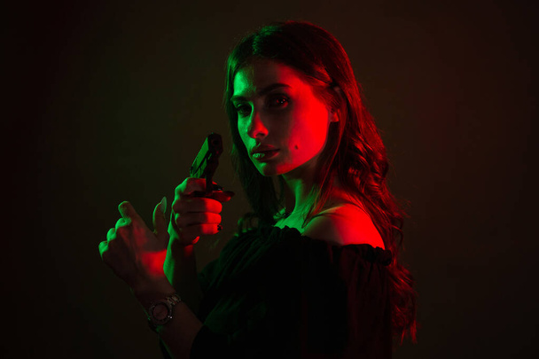 Красивая молодая женщина в темноте в клубе держит в руках пистолет. силуэт девушки в красно-зеленых цветах. Фонарь - Фото, изображение