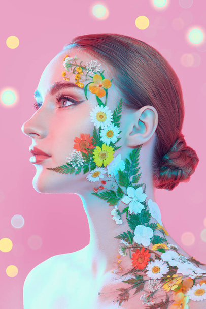 A beautiful red-haired young woman with elegant makeup and summer flowers on her face and shoulder posing in profile. Pink background with lights. Perfumery. Make-up and cosmetics. Beauty and flowers. - Valokuva, kuva