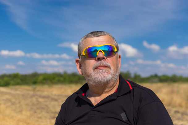 Outdoor portrait of a bearded senior in sunglasses against blue cloudy sky and agricultural field - Photo, Image