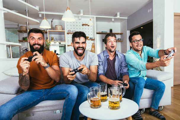 дружба, технологии и концепция отдыха - улыбающиеся друзья-мужчины с геймпадами и пивом, играющие дома в видеоигры  - Фото, изображение