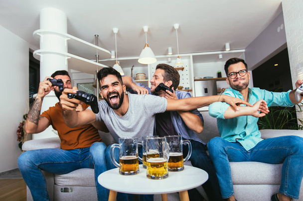 friendship, technology and leisure concept - smiling male friends with gamepads and beer playing video game at home  - Fotó, kép