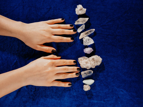Top view female hands with black nails lying on table with gem stones. Blue velvet for background. - 写真・画像