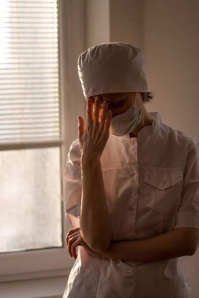 Eine gestresste Ärztin fasst sich an ihre Stirn und trägt eine Gesichtsmaske.   - Foto, Bild