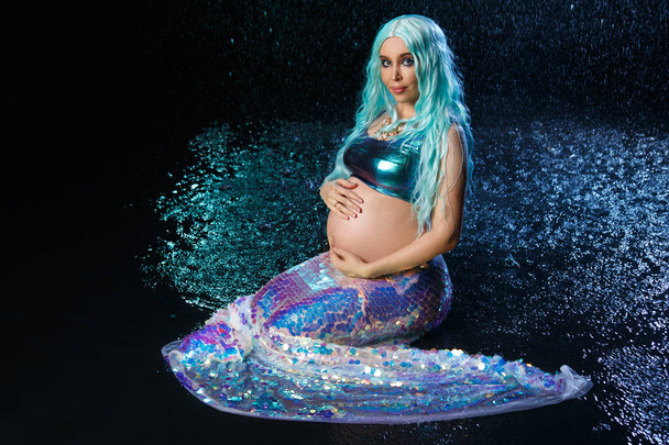 Portrait of a beautiful pregnant girl with a big belly in a mermaid costume with blue hair against a background of dark water. - Фото, изображение