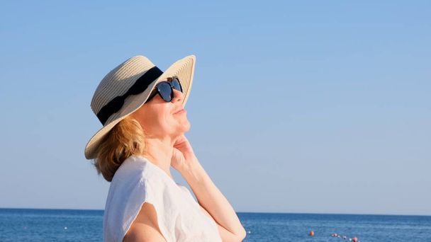Portrait of a contented 50 year old woman wearing a straw hat and sunglasses enjoying the sun against the blue sea. Summer, vacation, vacation, active retirees - Photo, Image