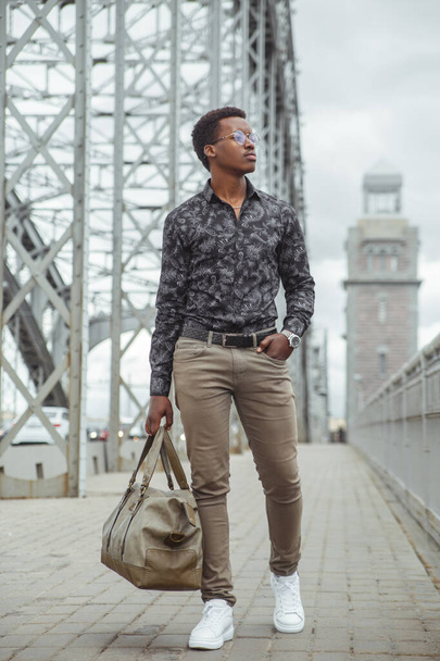 A young attractive black man with a large bag in his hand walks across the bridge in the city. - 写真・画像